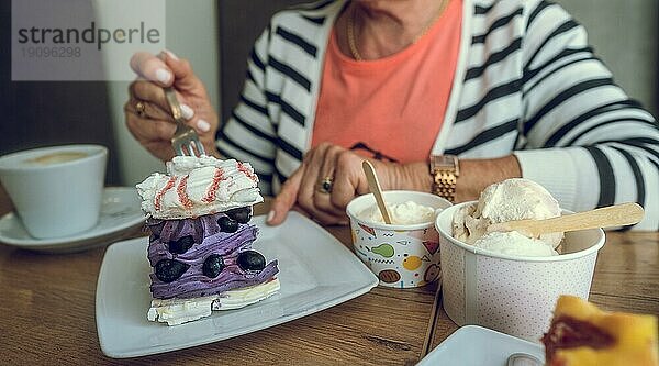 In einem Kaffeehaus. Lila Blaubeerkuchen  Eiscreme und Kaffee auf dem Tisch. Hand einer älteren Frau mit einer Kuchengabel. Kopierraum