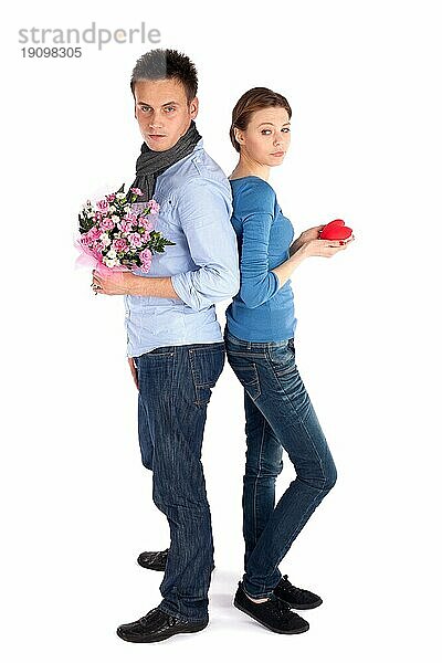 Ganzkörper junges attraktives Paar steht Rücken an Rücken  Frau hält Herz  Mann hält Blumen  vor weißem Hintergrund