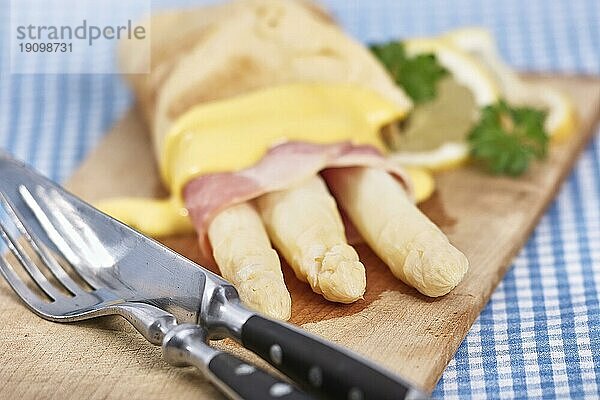 Weißer Spargel eingehüllt mit Schinken und Omlette liegt auf einem rustikalen Holzbrett. Links im Vordergrund liegt rustikales Besteck. Das Bild hat ein feines Bokeh