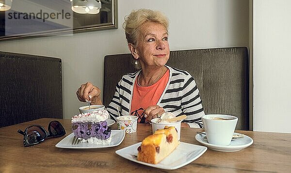 Attraktive  fröhliche ältere Frau trinkt Kaffee  während sie in einem Café sitzt. Porträt einer reifen Frau in einer Cafeteria. Bunte Torten und Kaffee auf dem Tisch. Angenehme entspannende Atmosphäre. Hübsche Frau mittleren Alters mit einer Tasse Kaffee