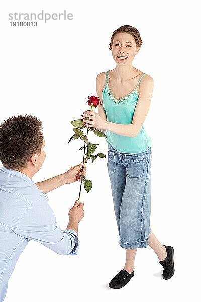 Mann überreicht einer glücklichen  fröhlichen jungen Frau eine einzelne rote Rose  vor weißem Hintergrund