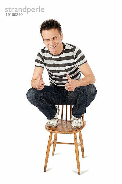 Lächelnd gut aussehend junger Mann sitzt auf Stuhl gestikulieren ok Zeichen isoliert gegen weißen Hintergrund