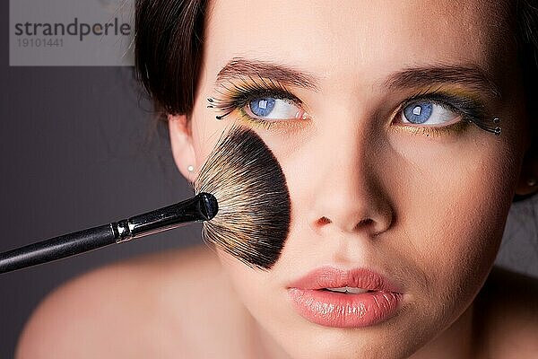 Beauty Shooting einer jungen Frau mit langen Wimpern  blaün Augen und Schminkpinsel  der ihr Gesicht berührt. Geringe Tiefenschärfe