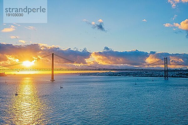 Blick auf die Brücke 25 de Abril  das berühmte touristische Wahrzeichen von Lissabon  die Lisboa und Almada auf der Halbinsel Setubal über den Fluss Tejo mit der Silhouette einer Touristenyacht bei Sonnenuntergang und einem fliegenden Flugzeug. Lissabon  Portugal  Europa