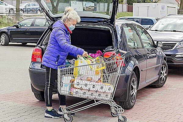 Eine reife Frau mit Schutzmaske lädt auf dem Parkplatz Einkäufe in ein Auto. Alltagsleben während einer Coronavirus Pandemie. Konzept Einkaufen