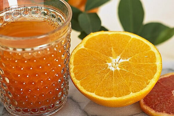 Neben einem Glas Saft liegt eine halbierte Apfelsine. Im Hintergrund sind weitere Früchte und Zweige. Das Bild hat ein feines Bokeh