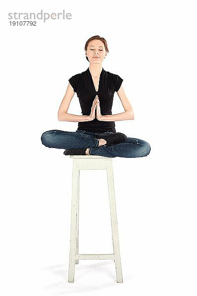 Schlanke Frau  die auf einem hohen Hocker sitzt und Yoga Meditationstechniken praktiziert  vor weißem Hintergrund