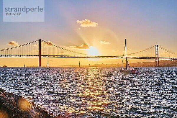 Blick auf die Brücke 25 de Abril  das berühmte touristische Wahrzeichen Lissabons  das Lisboa und Almada auf der Halbinsel Setubal verbindet  über den Fluss Tejo mit der Silhouette einer Touristenyacht bei Sonnenuntergang. Lissabon  Portugal  Europa