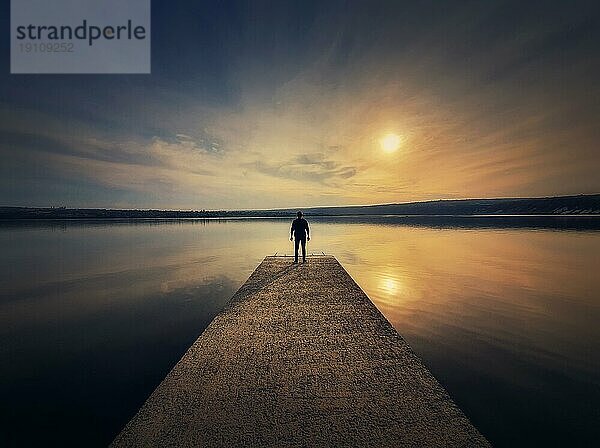 Mann  der allein auf dem Pier steht und den Sonnenuntergang betrachtet  der sich im ruhigen Wasser des Sees spiegelt. Scenic Ansicht mit einer einsamen Person auf dem Dock Blick auf den Horizont