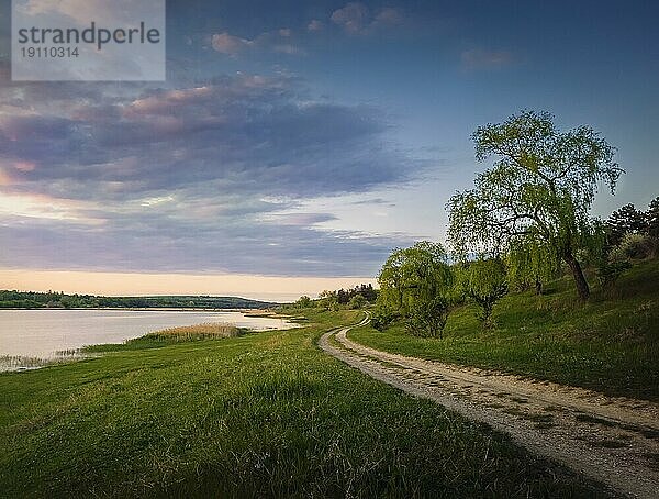 Ländliche Landschaft mit einer Landstraße zwischen See und Wald. Idyllische Sommerszene  friedlicher Abend mit buntem Sonnenuntergangshimmel