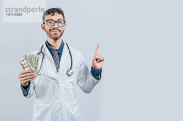 Schöner lächelnder Arzt zeigt Geld und zeigt auf die Seite. Glücklicher junger Arzt  der Geld hält und auf die Seite zeigt. Lateinischer Arzt