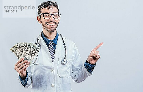 Glücklicher junger Arzt hält Geld und zeigt zur Seite. gutaussehender lächelnder Arzt zeigt Geld und zeigt auf die Seite. Lateinischer Arzt