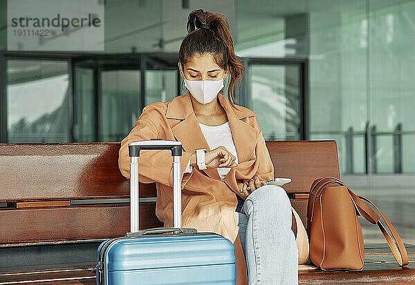 Frau mit medizinischer Maske  die während der Pandemie auf dem Flughafen auf ihre Uhr schaut