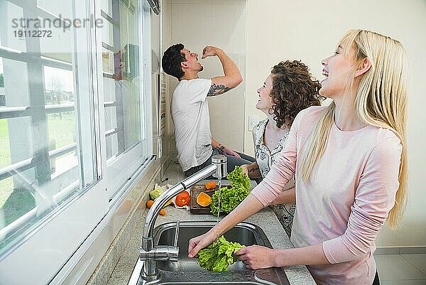 Zwei junge Frauen putzen lachend Salatgemüse  während ein Mann Karotten ißt