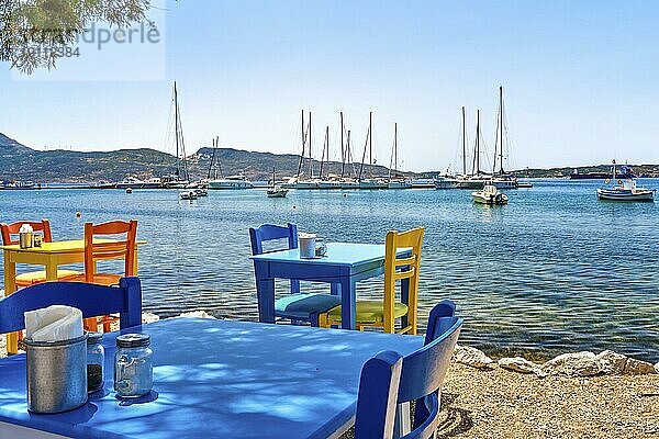 Bunte Stühle und Tische einer griechischen Taverne am Wasser an einem sonnigen Tag. Urlaub in Griechenland  Sommer  Restaurant  Essen im Freien  Hafen  Jachthafen  Yachten und Boote  Mittelmeer entspannen  Kykladen und Ägäischen Inseln. Selektiver Fokus  unscharfer Hintergrund
