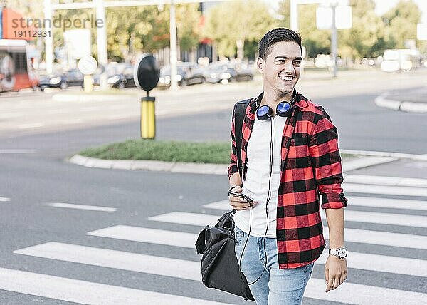 Glücklicher junger Mann mit Mobiltelefon beim Überqueren der Straße