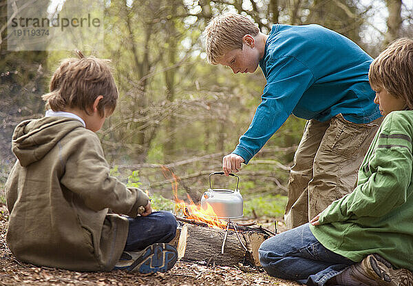 Drei Jungen am Lagerfeuer kochen Wasser in einem Wasserkocher