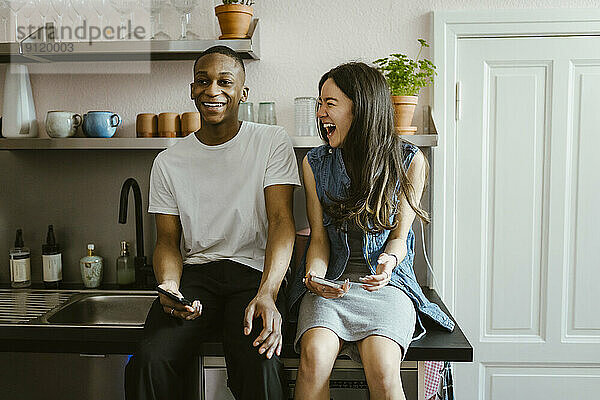 Fröhliche junge Freunde sitzen mit Smartphones auf dem Küchentisch zu Hause
