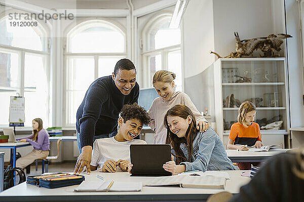 Glückliche männliche und weibliche Schüler  die sich beim Betrachten eines Tablet-PCs mit einem Lehrer im Klassenzimmer vergnügen