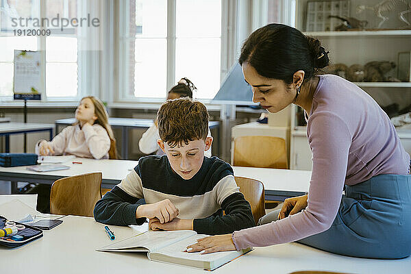 Eine Lehrerin hilft einem Schüler beim Lesen in einem Buch  während er auf dem Schreibtisch im Klassenzimmer sitzt