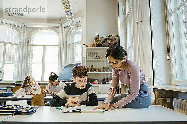 Eine Lehrerin hilft einem Schüler beim Lesen eines Buches  während er am Schreibtisch im Klassenzimmer sitzt