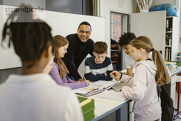 Glückliche Lehrerin mit multirassischen Schülern am Schreibtisch im Klassenzimmer