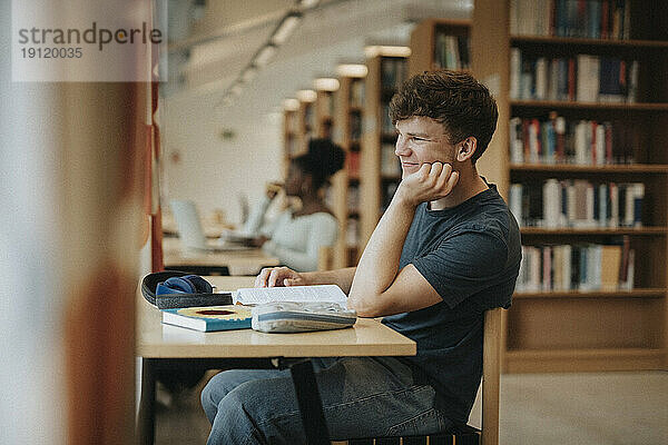 Seitenansicht eines lächelnden Studenten  der in einer Bibliothek sitzt und seine Hand auf das Kinn legt