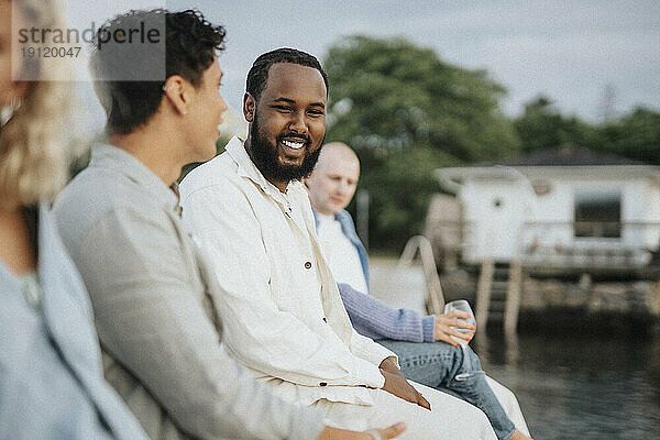 Lächelnder junger Mann im Gespräch mit einem männlichen Freund auf einem Steg in der Nähe eines Sees