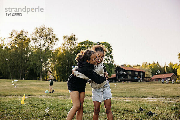 Glückliche Freundinnen umarmen sich gegenseitig  während sie auf dem Spielplatz im Gras stehen