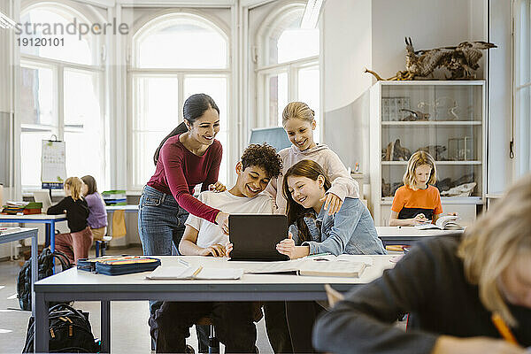 Lächelnde männliche und weibliche Schüler teilen sich einen Tablet-PC mit einem Lehrer im Klassenzimmer