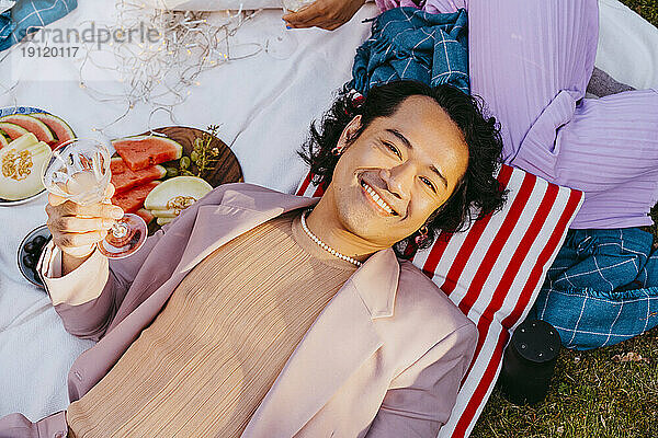 Hochwinkliges Porträt eines lächelnden schwulen Mannes  der ein Weinglas hält  während er sich im Hinterhof hinlegt