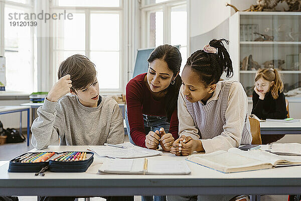 Lehrer diskutiert mit männlichen und weiblichen Schülern  die am Schreibtisch im Klassenzimmer sitzen