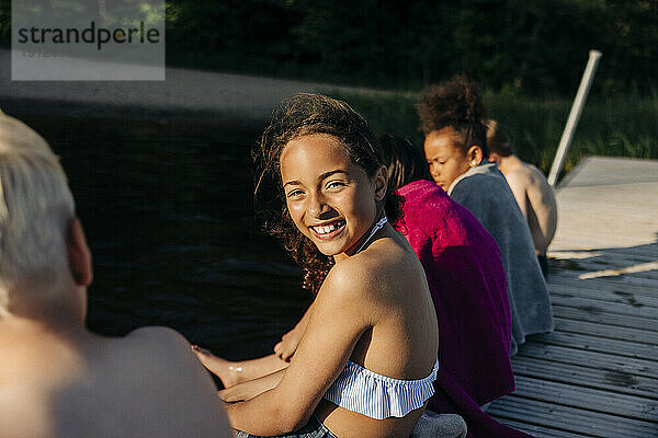 Seitenansicht Porträt von lächelnden Mädchen sitzen mit Freunden auf Steg in der Nähe von See im Sommerlager