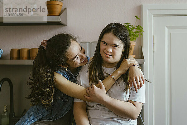 Glückliche junge Frau umarmt Schwester mit Down-Syndrom in der Küche zu Hause