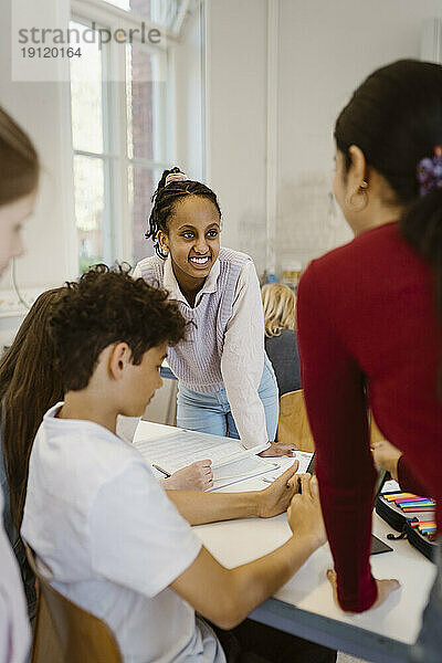 Lächelnder Schüler  der sich mit Freunden und Lehrer im Klassenzimmer an den Schreibtisch lehnt