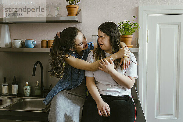 Lächelnde junge Frau umarmt Schwester mit Down-Syndrom in der Küche zu Hause
