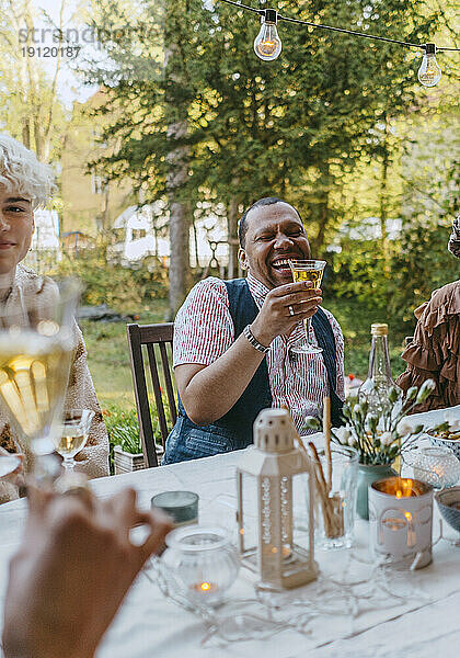 Glücklicher schwuler Mann  der ein Weinglas hält  während er mit Freunden bei einer Dinnerparty im Hinterhof sitzt