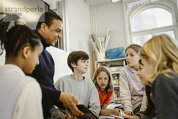 Männlicher Lehrer teilt sich einen Tablet-PC mit Schülern  die im Klassenzimmer diskutieren