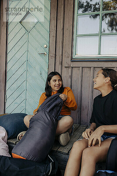 Glückliche Freundinnen sitzen mit Rucksäcken auf der Veranda vor der Hütte im Sommercamp