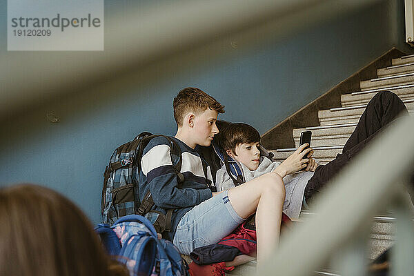 Männliche Freunde teilen sich ein Smartphone  während sie auf der Treppe eines Schulgebäudes sitzen