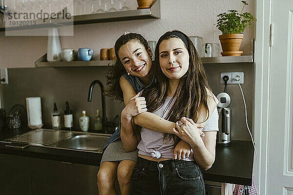 Porträt einer glücklichen Frau  die ihre Freundin von hinten umarmt  während sie zu Hause in der Küche sitzt