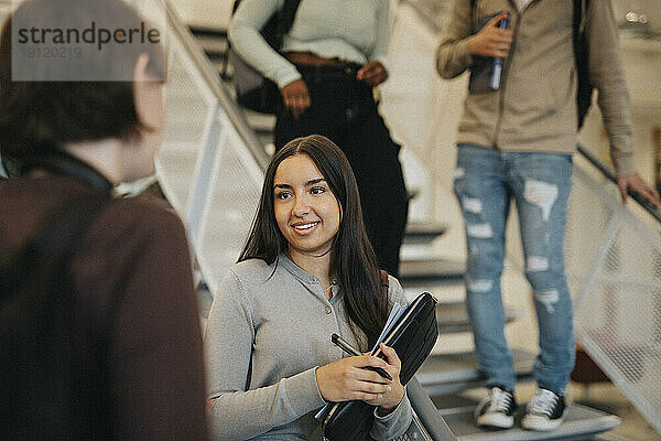 Lächelnde Studentin  die mit einem Freund diskutiert  während sie an einer Treppe in der Universität steht