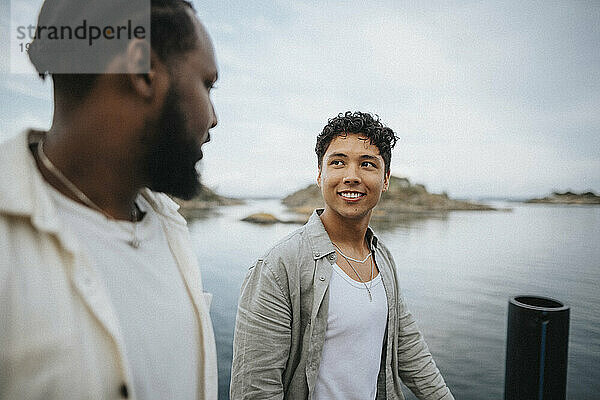 Lächelnder Mann  der einen männlichen Freund beim Spaziergang in der Nähe eines Sees ansieht