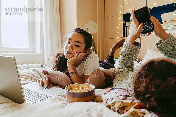 Junge Frau  die einen Laptop benutzt  während ihr Freund auf dem Bett eine Textnachricht über sein Smartphone sendet