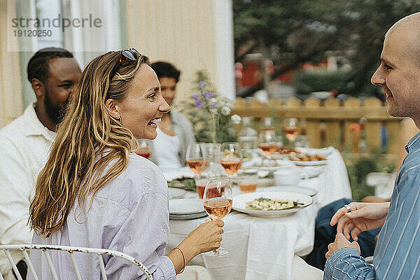 Glückliche junge Frau hält ein Weinglas in der Hand und lacht mit einem männlichen Freund während einer Dinnerparty im Café