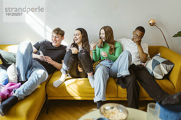 Fröhliche junge gemischtrassige Freunde benutzen ihr Smartphone  während sie zu Hause auf dem Sofa sitzen