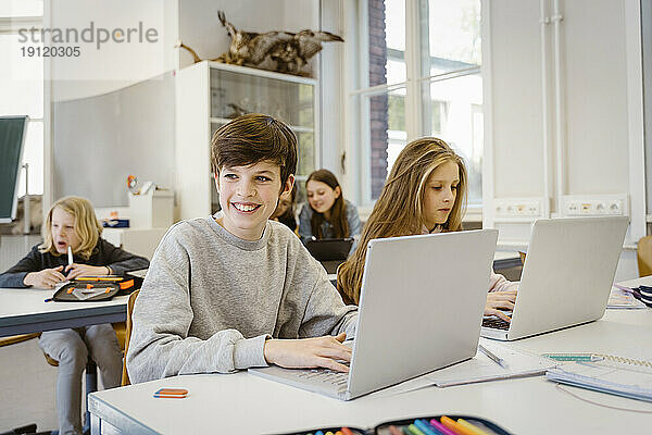 Glücklicher Junge  der wegschaut  während er mit seinem Laptop neben einer Freundin am Schreibtisch im Klassenzimmer sitzt