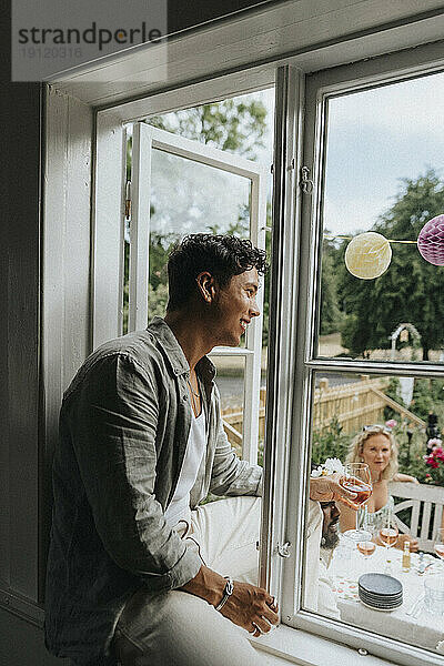 Lächelnder junger Mann  der auf der Fensterbank sitzt und seinen Freunden beim Feiern im Café zusieht