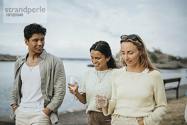 Mann im Gespräch mit Freundinnen  die Getränke halten  während er in der Nähe eines Sees spazieren geht