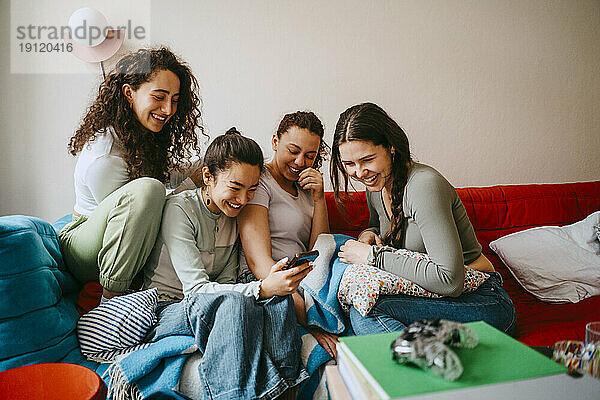 Glückliche junge Freundinnen schauen auf ihr Smartphone  während sie zu Hause auf dem Sofa sitzen